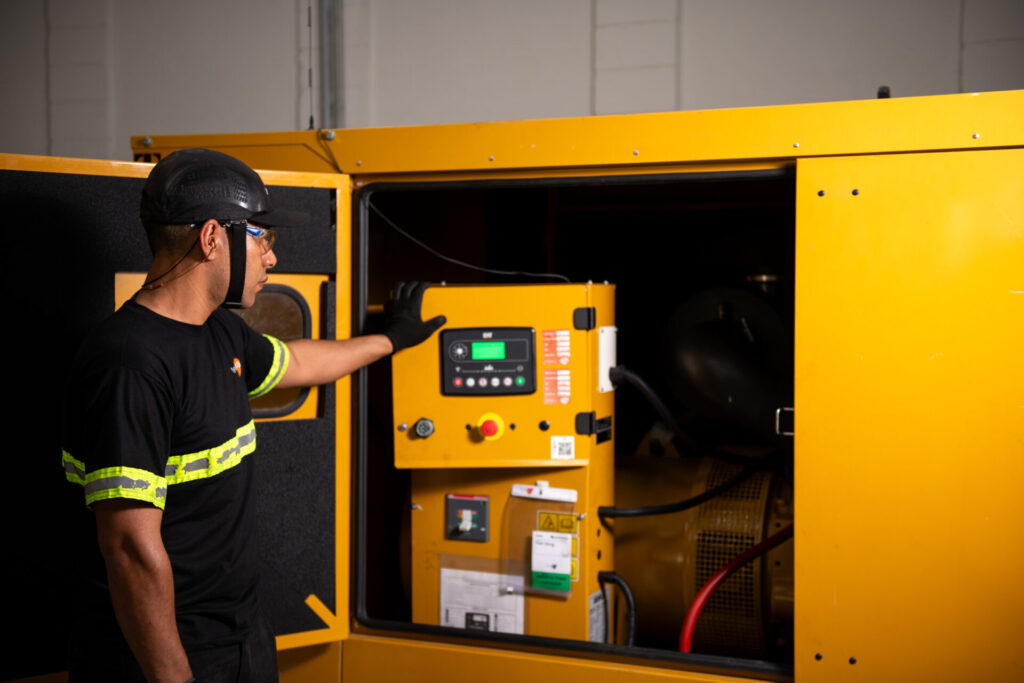 Profissional Tecnogera, com EPI completo, opera painel de gerador trifásico amarelo com compartimentos abertos no interior iluminado.