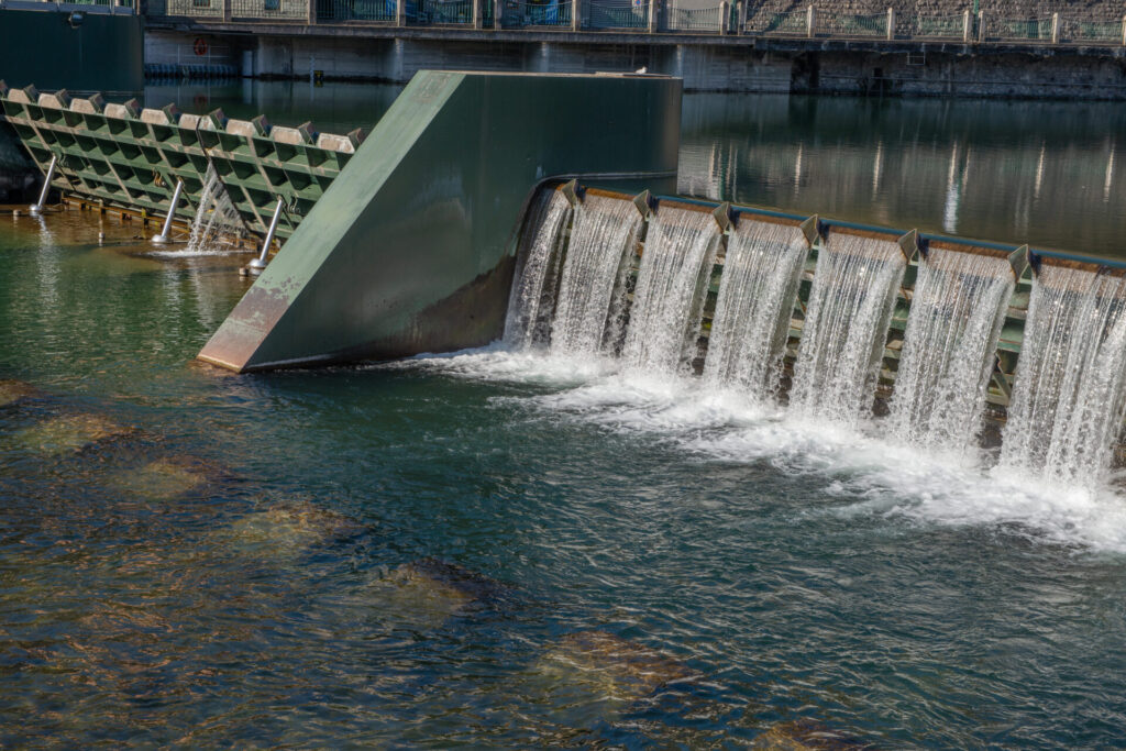 Uma comporta metálica moderna cruza o rio, controlando a água em degraus. Ao fundo, os prédios se refletem.