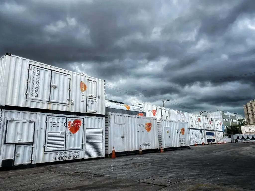 Diversos geradores brancos da Tecnogera, estão alinhados em um pátio asfaltado, prontos para fornecer energia temporária. Cones de sinalização laranja demarcam a área enquanto nuvens escuras anunciam chuva e prédios altos aparecem suavemente ao fundo