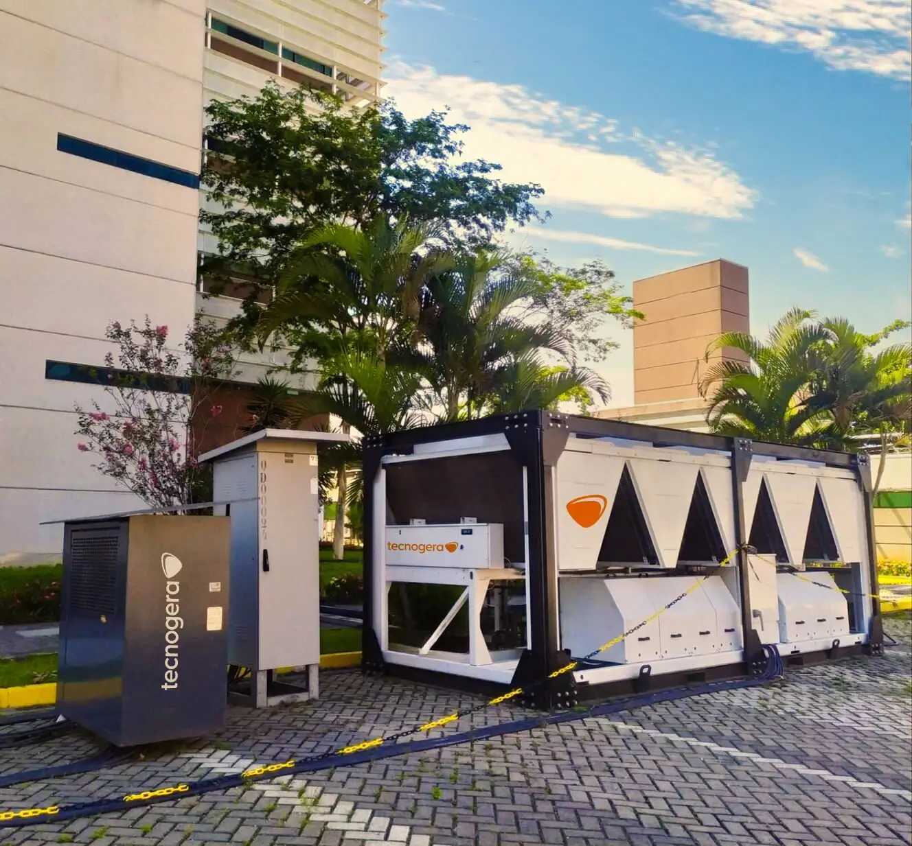 Um grande chiller branco está instalado ao ar livre, sobre um piso pavimentado próximo a um prédio moderno, em meio a árvores e plantas. O local está sinalizado com fita zebrada amarela e preta, destacando o robusto sistema de refrigeração da Tecnogera sob o céu azul