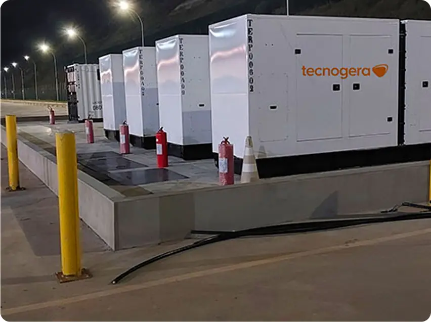 Vários geradores brancos com o logotipo da Tecnogera estão posicionados sobre uma plataforma de concreto, representando suas soluções em energia. Perto deles, há balizadores amarelos, extintores de incêndio e um cone de trânsito. À noite, a área fica iluminada pelos postes de luz.