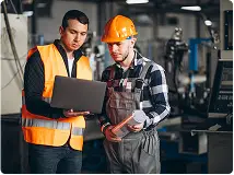 Na imagem, dois homens em uma fábrica conversam sobre soluções em energia enquanto analisam um laptop. Um deles usa colete de segurança laranja e o outro, de capacete e macacão, segura uma planta baixa. Ao fundo, é possível ver máquinas e equipamentos industriais da Tecnogera.