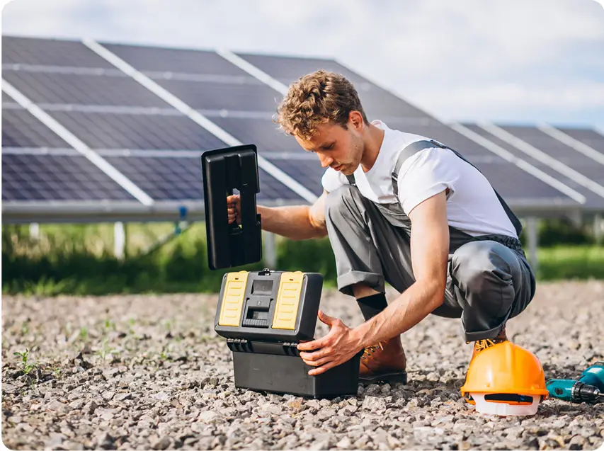 Um colaborador da Tecnogera, usando macacão cinza, está ajoelhado sobre o cascalho e abrindo uma caixa de ferramentas preta e amarela próxima a grandes painéis solares - mostrando as soluções em energia da empresa. Ao lado dele, repousa um capacete laranja no chão. O cenário conta ainda com grama verde e um céu parcialmente nublado ao fundo.