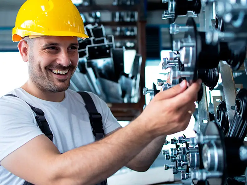 Um homem sorridente, usando capacete amarelo e camisa branca, ajusta peças metálicas de uma máquina em um ambiente industrial. Ele parece ser um engenheiro especialista em soluções de energia da Tecnogera, trabalhando entre diversos componentes mecânicos e equipamentos.