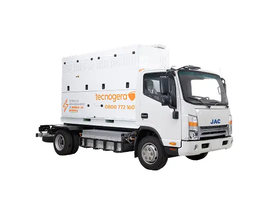 Um caminhão branco da JAC traz uma grande bateria instalada na carroceria. O equipamento exibe a marca Tecnogera com seu logotipo laranja e telefone para contato, destacando as soluções em energia oferecidas pela empresa. A imagem mostra o caminhão isolado sobre um fundo branco, visto de um ângulo frontal lateral.