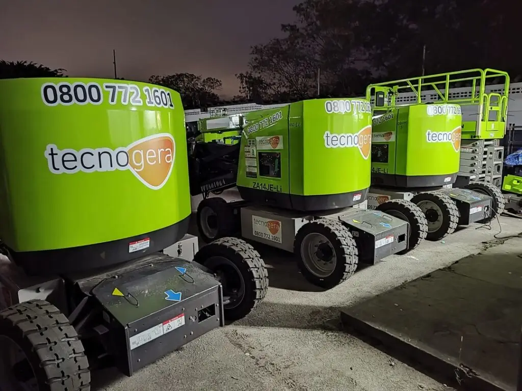 Quatro plataformas elevatórias móveis, pintadas de verde vibrante e com o logotipo e telefone da Tecnogera, estão estacionadas lado a lado sobre um piso de concreto à noite, prontas para realizar a limpeza da fachada. Ao fundo, aparecem algumas árvores sob o céu escuro.