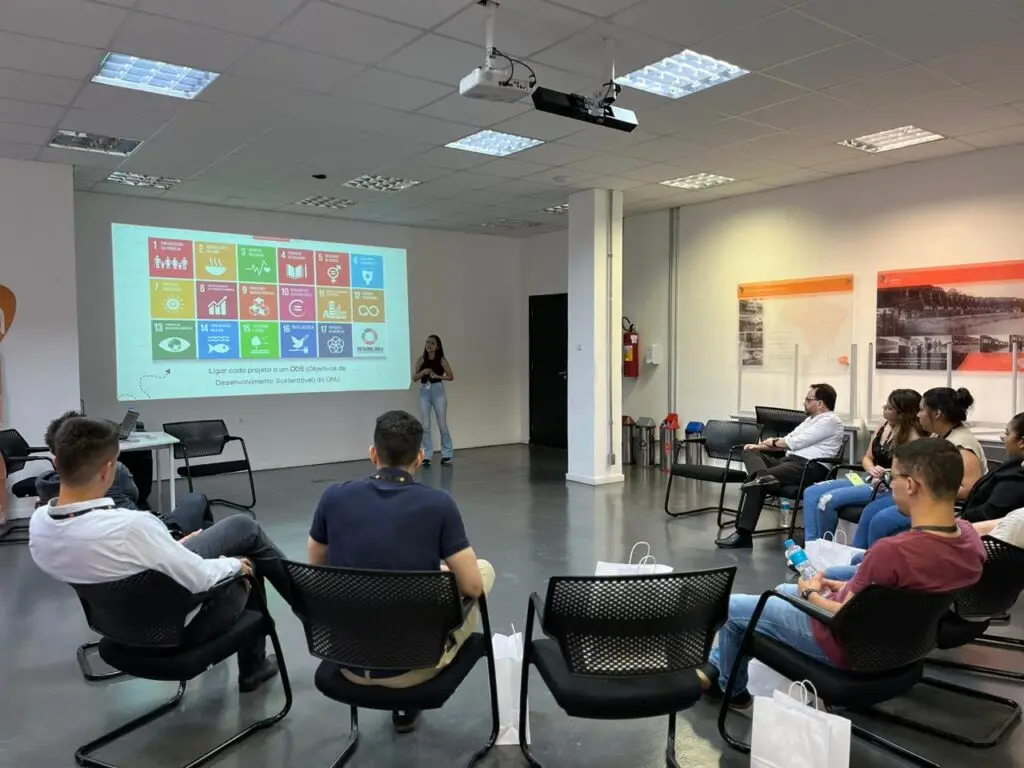 Na imagem, uma mulher se apresenta diante de uma tela com ícones coloridos e textos para um grupo sentado em um ambiente moderno e bem iluminado. O espaço conta com cadeiras pretas, projetor, sacolas brancas, pôsteres do GerAção nas paredes e destaca o tema "futuro". A cena faz parte das iniciativas da Tecnogera.