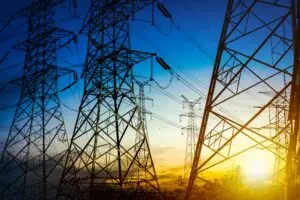 Torres de energia metálicas e linhas de transmissão aparecem em silhueta diante de um pôr do sol colorido, com o sol se pondo atrás das árvores. Esse é um cenário típico do trabalho das concessionárias de energia ao monitorar índices como DEC e FEC para garantir a qualidade do serviço - algo que faz parte das soluções oferecidas pela Tecnogera.
