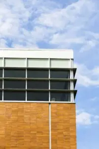 Um prédio moderno com fachada composta por painéis verticais de madeira na parte inferior e grandes janelas de vidro escuro emolduradas em branco na parte superior, contrastando com um céu azul pontuado por nuvens. Um cenário perfeito para a Tecnogera realizar a limpeza da fachada.