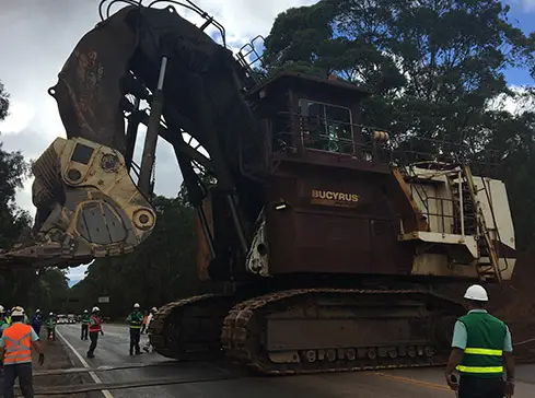 Uma escavadeira Bucyrus de grande porte, com trilhos e caçamba levantada, está parada em uma estrada em um local de mineração. Perto dela, alguns trabalhadores usando coletes de segurança e capacetes acompanham as atividades. Ao fundo, é possível ver árvores e um céu nublado, ilustrando o ambiente operacional da Tecnogera.
