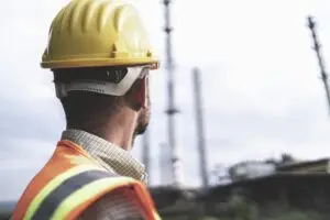 Um homem com capacete amarelo e colete refletivo laranja observa, ao ar livre, estruturas industriais ao longe, ressaltando a importância da NR-35 e da segurança do trabalho em obras. A Tecnogera está sempre atenta a esses cuidados em seus projetos.