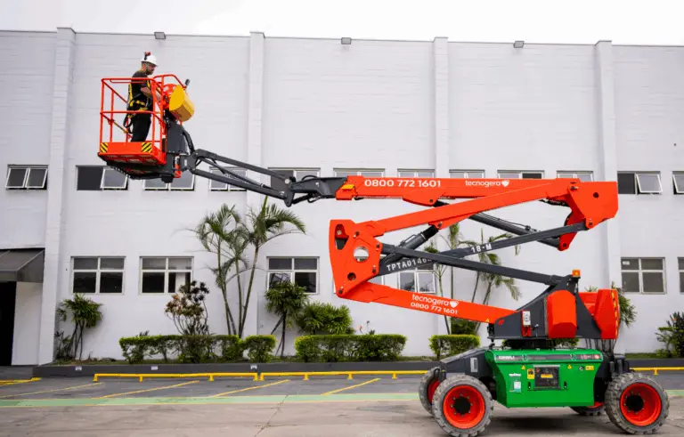 Uma pessoa equipada com itens de segurança está na cesta elevada de uma plataforma articulada laranja e verde, utilizada para trabalhos em altura pela Tecnogera. Ao fundo, é possível ver um prédio industrial branco, com grandes janelas e palmeiras na frente.