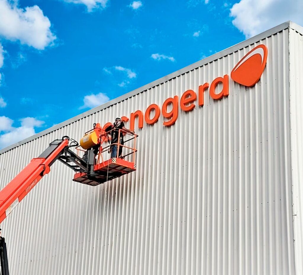 Dois colaboradores utilizam uma plataforma elevatória para instalar ou fazer a manutenção de uma placa laranja com o nome "nogera" na fachada ondulada de um grande prédio industrial, sob um céu azul e ensolarado. Tecnogera: muito mais do que locação de geradores.