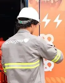 Uma pessoa com capacete branco e jaqueta cinza, que traz o nome Tecnogera nas costas, observa um veículo laranja identificado por símbolos de raio branco. A jaqueta possui faixas amarelas refletivas nas mangas.