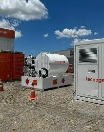 Cenário industrial ao ar livre, com equipamentos brancos e vermelhos identificados com a marca Tecnogera posicionados sobre um piso pavimentado. O local conta com um tanque de combustível, geradores, um contêiner laranja e cones de segurança. O céu é azul e há algumas nuvens espalhadas.