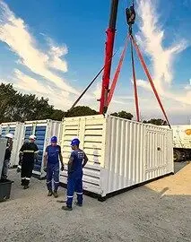 Uma grua ergue um grande contêiner branco com cintas vermelhas em um canteiro de obras. Três trabalhadores, usando uniforme azul e capacete, acompanham de perto a movimentação, enquanto outra pessoa equipada para segurança passa pelo local. Ao fundo, é possível ver outros contêineres semelhantes e algumas árvores. Essa cena representa bem as soluções oferecidas pela Tecnogera em ambientes industriais e de construção civil.