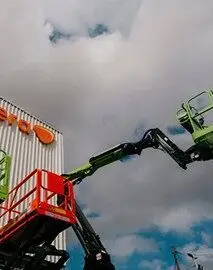 Dois elevadores do tipo boom lift, um verde e outro vermelho, estão posicionados e estendidos em direção a um prédio branco de estrutura ondulada, marcado por letras e formas na cor laranja. O céu nublado com grandes nuvens ao fundo cria um cenário imponente. Essa imagem destaca a atuação da Tecnogera em soluções para trabalho em altura.