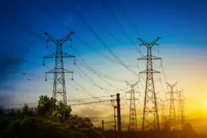 Na imagem, várias torres metálicas de energia elétrica, se estendem por uma paisagem repleta de árvores verdes, ilustrando os desafios e soluções envolvidos na geração de energia. Ao fundo, o céu muda do azul intenso para tons alaranjados e amarelos típicos do amanhecer ou entardecer.