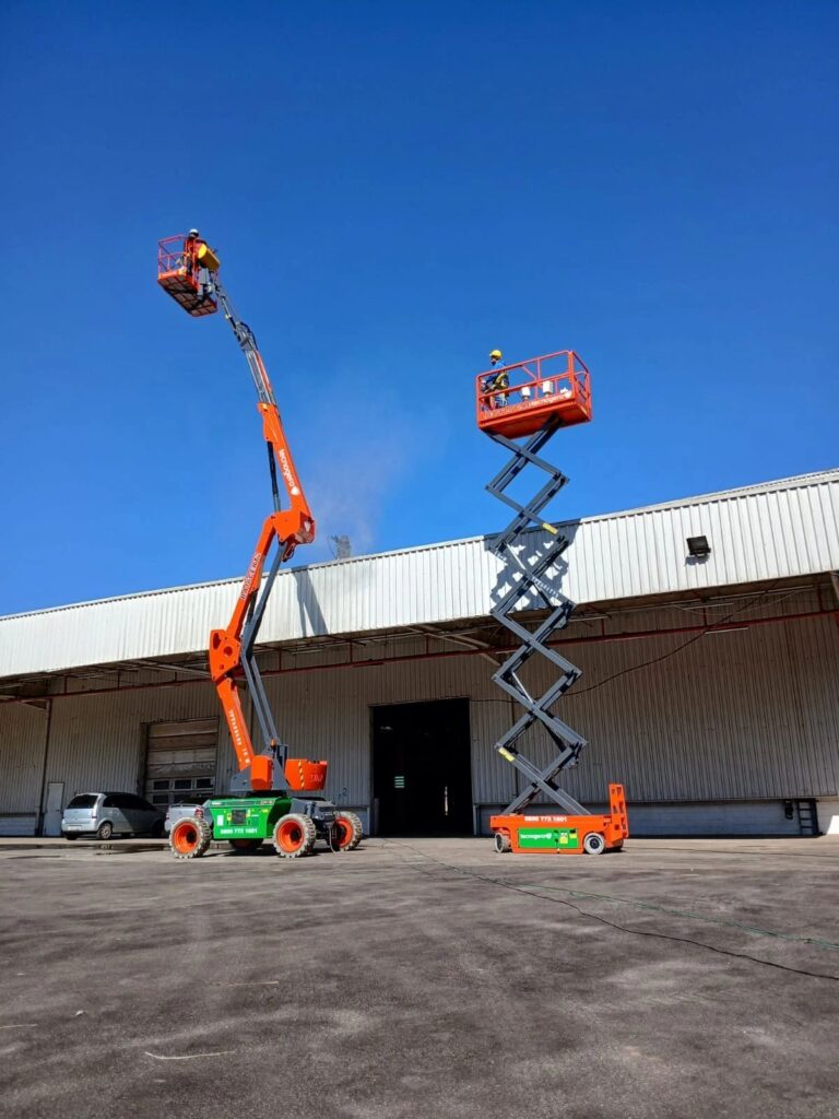 Dois operadores profissionais de plataformas elevatórias da Tecnogera trabalham em frente a um galpão industrial. Ao fundo, é possível ver o céu azul.