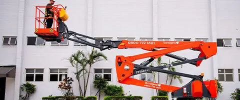 Colaborador Tecnogera com EPI opera plataforma elevatória laranja ao lado de prédio branco com janelas retangulares e plantas verdes.
