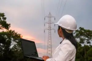 Profissional com notebook e capacete analisa torre de energia entre árvores ao pôr do sol, mostrando atuação em campo.