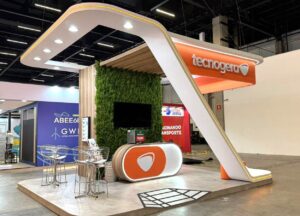 Stand da Tecnogera na Brazil Windpower 2025 com tons branco e laranja, estrutura arco, painel verde e balcão personalizado.