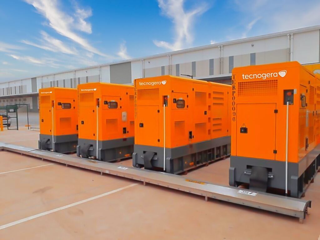 Quatro geradores laranja da Tecnogera, alinhados e prontos para uso em emergências, ao lado de um galpão moderno.