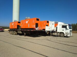 O Caminhão branco Tecnogera transporta quatro geradores laranja sobre o pátio, com base de torre e árvores ao fundo.