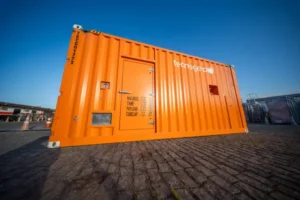 Container laranja da Tecnogera em piso de paralelepípedo, sob céu azul. Possui porta, respiros e avisos. Outros itens ao fundo.