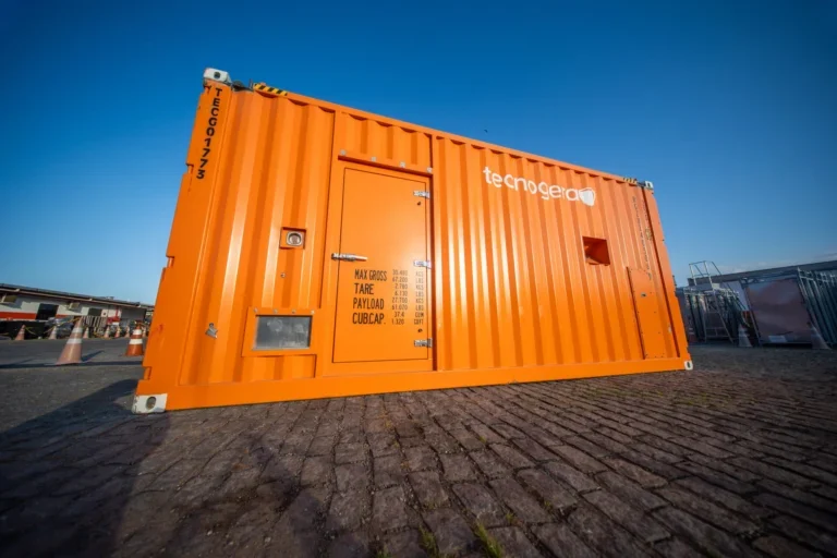 Container laranja da Tecnogera em piso de paralelepípedo, sob céu azul. Possui porta, respiros e avisos. Outros itens ao fundo.