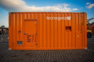 Gerador laranja da Tecnogera sobre paralelepípedos, com porta, respiros e ventilações. Céu azul ao fundo.