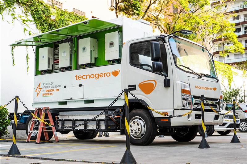 A Tecnogera estacionou seu caminhão branco com o painel lateral aberto, mostrando equipamentos elétricos, próximo a árvores e prédios.