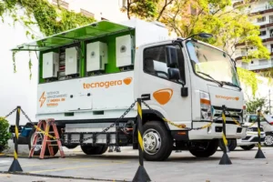 Um caminhão da Tecnogera com unidades BESS está estacionado em um terreno, próximo a correntes e postes de amarração, com plantas e edifícios ao fundo.