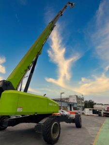 Plataforma Elevatória Tecnogera de 51m, verde, braço estendido, em pátio industrial sob céu azul; carros e prédios ao fundo.