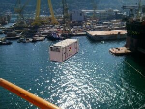 Tecnogera: Um guindaste levanta um contêiner branco sobre a água, com a luz do sol e estruturas industriais ao fundo.