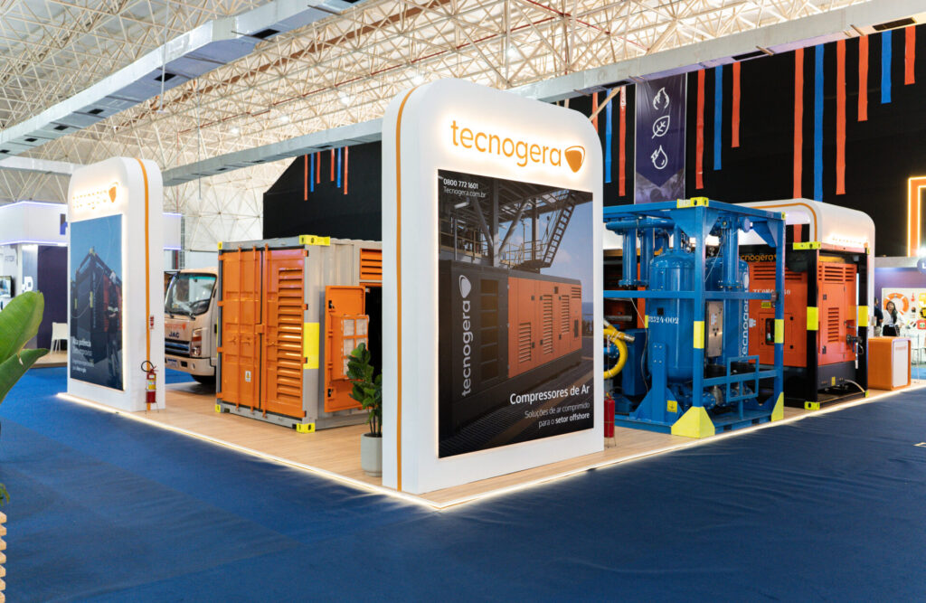 Estande da Tecnogera em Macaé Energy, com equipamentos para offshore laranja sobre piso de madeira e tapete azul ao redor.