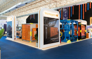 Estande da Tecnogera em Macaé Energy, com equipamentos para offshore laranja sobre piso de madeira e tapete azul ao redor.