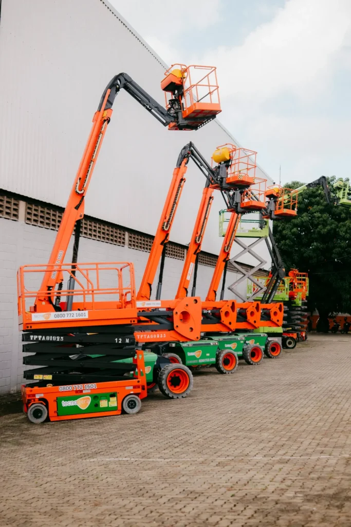 Plataformas elevatórias Tecnogera laranja e verde, braços erguidos ao lado do prédio branco. Solução prática sob céu nublado.