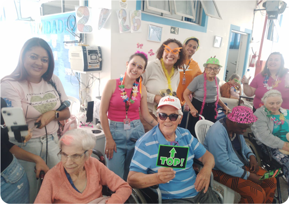 Colaboradores da Tecnogera e idosos posam sorrindo em um evento de responsabilidade social, com acessórios festivos e decoração alegre.