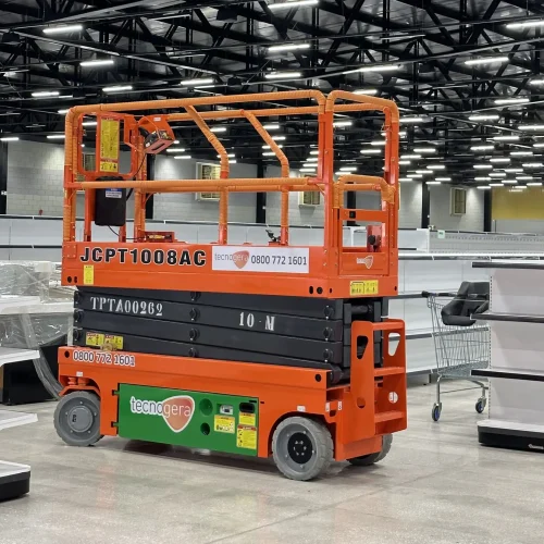 Uma plataforma elevatória tesoura na cor laranja vibrante, modelo "JCPT1008AC", chama a atenção no amplo espaço interno bem iluminado, com vigas pretas aparentes. Ao redor, prateleiras e estruturas sugerem que o local está em processo de instalação ou organização. A Tecnogera oferece soluções como essa para facilitar operações em altura com segurança e eficiência.