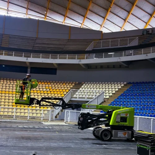 Profissional com capacete na plataforma PTA verde Tecnogera, trabalhando no alto de arena coberta, piso protegido por lonas pretas.