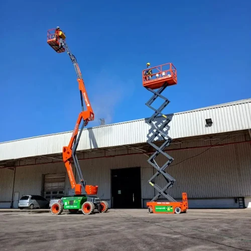 Dois profissionais da Tecnogera usam plataformas elevatórias - uma articulada laranja e outra tesoura verde-laranja - diante de um galpão.