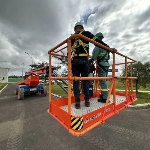 Dois trabalhadores, equipados com cintos de segurança, estão sobre uma plataforma elevatória laranja identificada com a inscrição "CAPACIDADE 230kg", em uma área externa asfaltada. A plataforma está suspensa e, ao fundo, é possível ver um grande equipamento do tipo boom lift sob um céu nublado. Essa é uma das soluções oferecidas pela Tecnogera para trabalho seguro em altura.