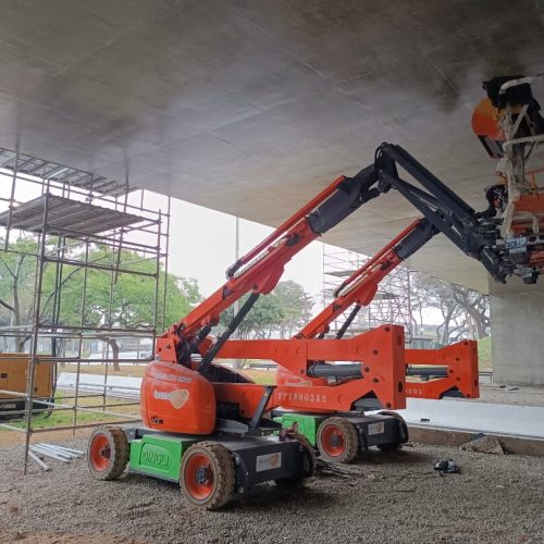 Dois elevadores articulados vermelhos da Tecnogera estão posicionados sob uma ponte de concreto, auxiliando na manutenção predial com segurança e eficiência. Um dos equipamentos está estendido para cima, permitindo que um colaborador alcance a parte inferior da estrutura, enquanto andaimes e árvores completam o cenário sobre o solo de cascalho.