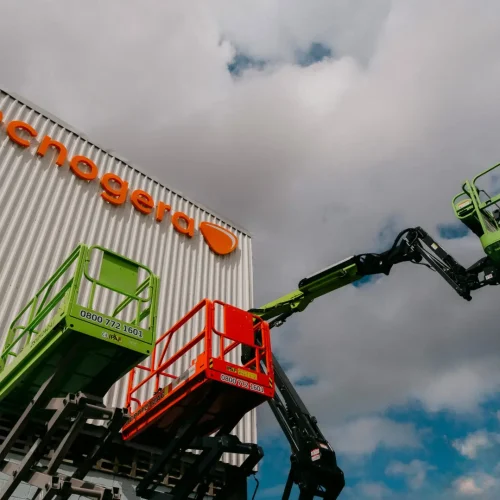 Duas plataformas elevadas, uma vermelha e outra verde, em um prédio de metal com a placa laranja da Tecnogera. Céu parcialmente nublado.