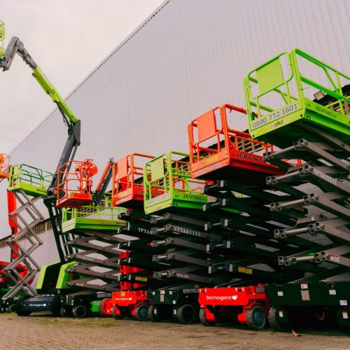 Uma fileira de plataformas para trabalhos em altura da Tecnogera, como tesouras e braços articulados em cores vibrantes - vermelho, verde e laranja - está posicionada em frente a um grande galpão. Um dos equipamentos, um braço articulado verde, aparece totalmente estendido com um colaborador trabalhando nas alturas sob o céu nublado.