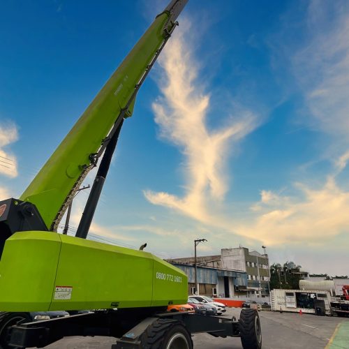 Plataforma elevatória verde da Tecnogera se destaca em altura, sob céu azul, ao lado de veículos e galpões industriais.