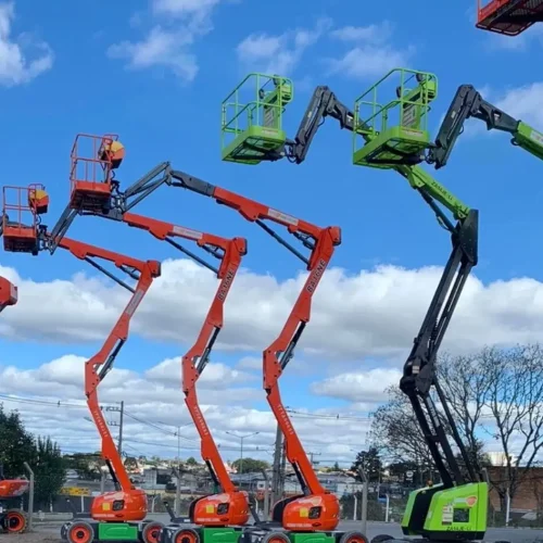 Plataformas elevatórias posicionadas em um pátio de cascalho sob um céu azul com nuvens, tendo ao fundo alguns prédios e árvores. Tudo isso faz parte da estrutura que a Tecnogera oferece para soluções eficientes em trabalhos em altura